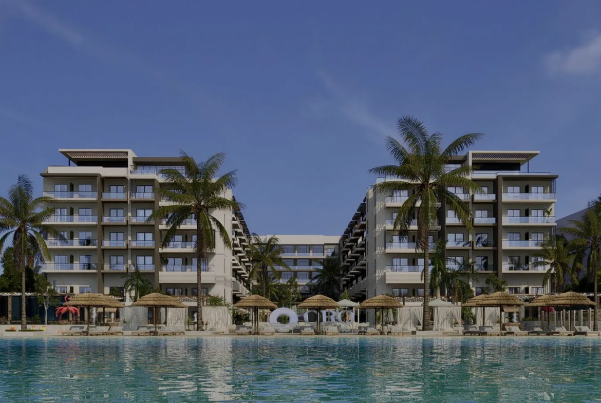 oro — developer visualisation — Modern beachfront resort with two six-story buildings, palm trees, thatched umbrellas, lounge chairs, and a large swimming pool in the foreground.