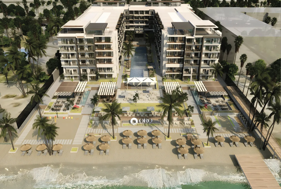oro — developer visualisation — Aerial view of a beachfront resort featuring two six-story buildings flanking a central pool, sandy beach with umbrellas and lounge chairs, palm trees, and a wooden pier extending into the ocean.