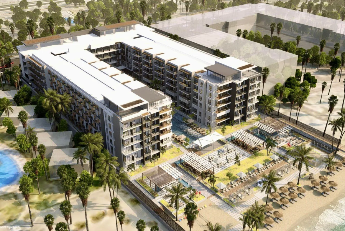 oro — developer visualisation — Aerial view of a modern beachfront resort with apartment buildings, a swimming pool, palm trees, shaded lounge areas, and beach umbrellas along the shoreline.
