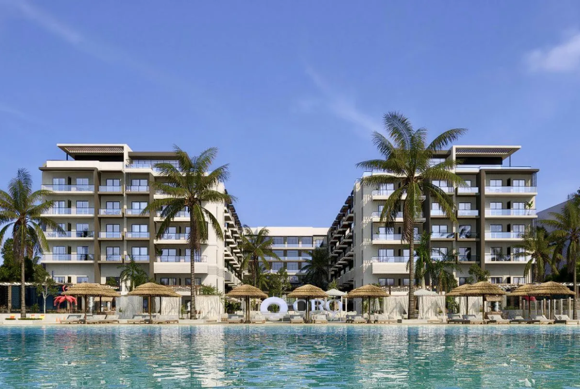 oro — developer visualisation — Modern resort buildings with balconies facing a large turquoise pool, surrounded by palm trees and thatched umbrellas under a clear blue sky.